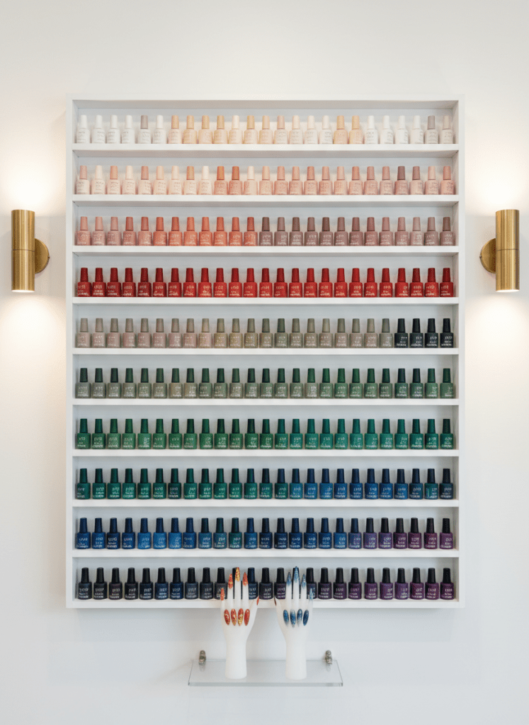 An atmospheric, photographic shot of a minimalist wall-mounted nail color display in a boutique studio, filled with rows of carefully arranged gel polish bottles in a gradient from soft neutrals to deep jewel tones. Below, a small glass shelf holds two silicone display hands showcasing seasonal themed nail sets: one with warm terracotta tones and delicate gold leaf, the other with cool smoky blues and subtle glitter ombré. Warm, diffused wall sconces cast a gentle glow, creating soft reflections on the bottles and adding depth to the scene. Captured straight-on with sharp focus throughout, the mood is curated and upscale, presenting the breadth of creative options offered by a skilled Valdese, NC nail technician.
