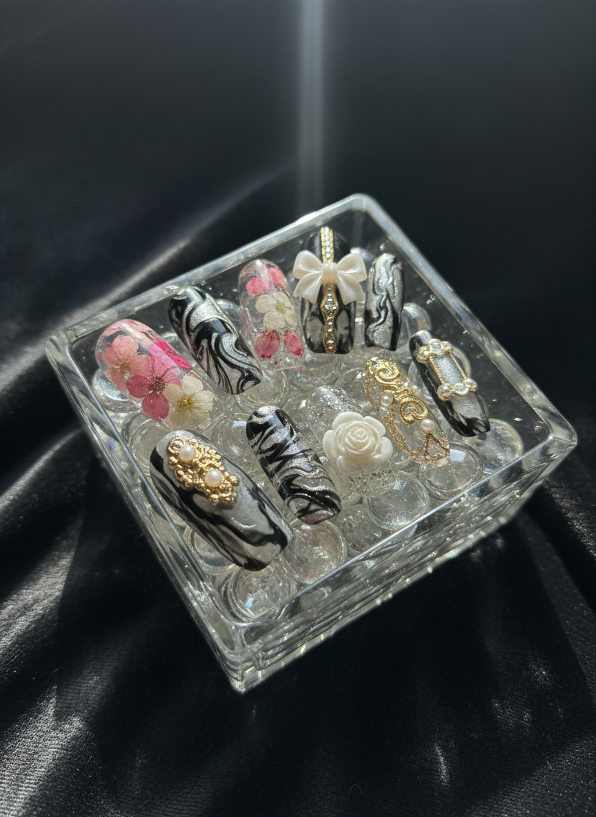 A dramatic photographic composition of a crystal-clear glass dish filled with assorted custom nail art tips, each resting like jewelry on a bed of tiny glass beads. Designs range from soft pink encapsulated florals to bold black-and-silver marble, with several nails featuring intricate 3D sculpted elements like tiny bows and petals. The dish sits on a velvety charcoal fabric, adding depth and contrast. A single focused spotlight from above creates sparkling highlights and rich shadows, making the nails appear like luxury accessories. Captured from close range with a slightly tilted angle and shallow depth of field, the ambiance is glamorous and artistic, perfectly suited for a sophisticated nail technician’s portfolio imagery.